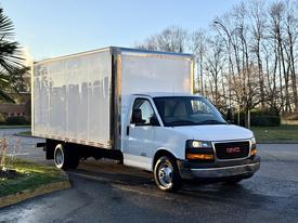 A white 2015 GMC Savana box truck with a flatbed and a large cargo area is parked at an angle with its headlights on
