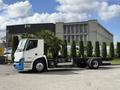 A 2022 Lion Lion6 electric truck chassis with a white and blue exterior standing in front of tall green hedges and a building
