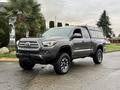 A gray 2016 Toyota Tacoma pickup truck with a silver canopy on the bed and black alloy wheels is parked on a wet street