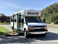 A 2014 Chevrolet Express shuttle bus with a white exterior and a yellow wheelchair ramp extended from the side door