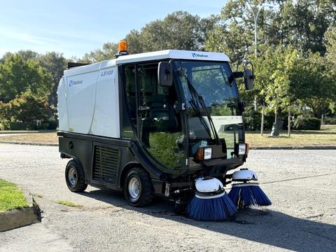 A 2017 Madvac LS100 street sweeper with dual front brushes and an orange flashing light on top parked on a street