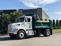 A white 2015 Peterbilt 337 dump truck with a green bed is parked with its bed raised, displaying wooden planks inside