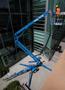 A blue 2016 Genie TZ-34/20 aerial lift with an extended platform and operator in a safety harness working at height near a building wall