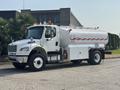 A 2010 Freightliner M2 106 tanker truck with a white body and red detailing equipped with multiple safety lights and large tank mounted on the back