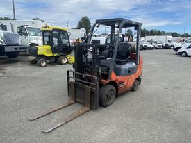 A 2010 Toyota 7FGGU25 forklift with an orange and black exterior is positioned in the foreground with forks lowered and a driver's seat visible