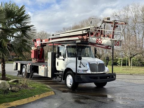 A 2010 International 4300 truck equipped with an aerial lift feature and a flatbed, parked on a street with a palm tree nearby