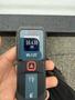 A hand holding a Bosch Blaze laser distance measurer displaying a reading of 10.430 feet
