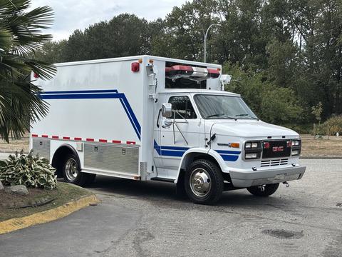 A white 1994 GMC Vandura 3500 with blue stripes and emergency lights parked at an angle showing its boxy shape and large wheels