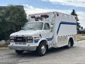 A 1994 GMC Vandura 3500 in white with blue stripes equipped as an ambulance with a light bar on top and large rear doors