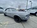 A 1968 Ford Mustang two door convertible in silver with a black soft top and black wheels parked among other vehicles