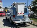 A blue 2006 Ford Econoline van with a white mounted utility box, ladder rack, and rear step bumper visible in the foreground