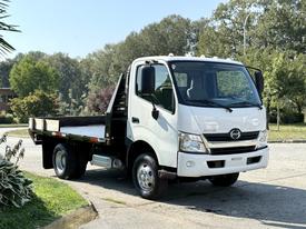 A white 2012 Hino 155 flatbed truck parked with a cabin featuring a gray interior and a flat load area at the back