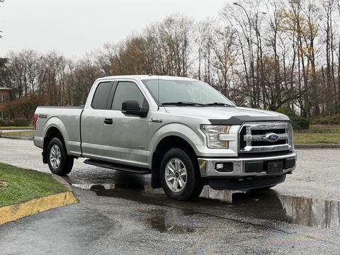 A silver 2015 Ford F-150 with a crew cab and chrome accents is parked on a wet surface with its front angled towards the viewer