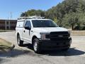 A white 2019 Ford F-150 with a ladder rack on top parked on a gravel surface with a distinctive grille and headlights
