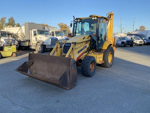 A yellow 2007 Komatsu WB156PS-5 backhoe loader with a large front bucket and large rear stabilizers parked at a construction site