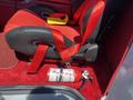 A black and red racing seat with a five-point harness and a fire extinguisher placed beside it on a red carpeted floor in a vehicle