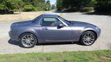 A 2007 Mazda Miata in gray, featuring a compact design with a hardtop, sporty wheels, and smooth lines, parked on a driveway