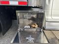 A silver metal storage box mounted on a truck with a strap and a metal piece inside, positioned next to a 2013 RAM 5500