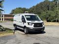 A white 2016 Ford Transit van parked with a ladder rack on top and side doors visible on a paved surface