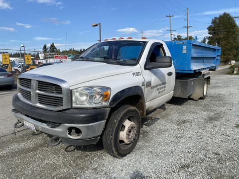 A white 2009 Dodge Ram 5500 truck with a blue flatbed is parked and facing left with visible dirt on its surface and a business logo on the door