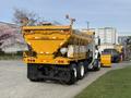 A yellow 2016 International 7600 dump truck with a snow plow attached at the front and a storage box at the rear