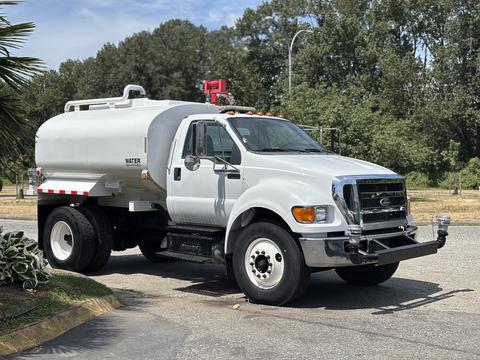A 2015 Ford F-750 water truck with a white tank body and large wheels parked on a street