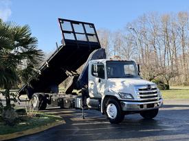 A 2015 Hino 358 truck with a raised dump bed, featuring a white cab, black dump body, and chrome details, positioned in a parking area