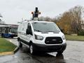 A 2019 Ford Transit van with a ladder rack on top parked in a wet area with a bus visible in the background