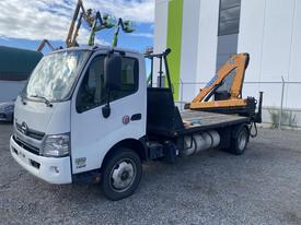 A 2019 Hino 195 flatbed truck with a yellow crane attached on the side parked on gravel
