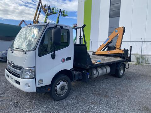 A 2019 Hino 195 flatbed truck with a yellow crane attached on the side parked on gravel