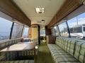 Interior view of a 1976 GMC Motorhome featuring plaid seating, a wooden dining table, kitchen area, and large windows allowing light into the space