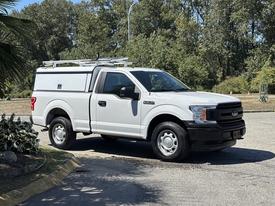 A white 2019 Ford F-150 is parked with a utility cap and a ladder rack on its roof, featuring a black grille and wheels
