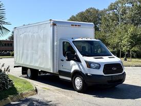 A 2015 Ford Transit box truck with a white body and black front bumper parked on a street