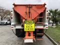 A 2012 International 7400 truck with an orange snowplow attachment and a bright yellow caution sign that reads "CAUTION STAY BACK 15 METERS" hanging from the rear