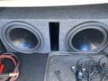 A pair of large black subwoofers enclosed in a gray carpeted box located in the trunk of a 2014 Mercedes-Benz CLA-Class with various cables nearby
