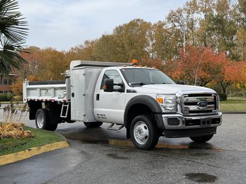 A white 2012 Ford F-550 truck with a flatbed and a metallic storage box on the side is parked on a wet pavement