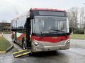 A 2016 Grande West Vicinity bus with a red and silver design features a lowered ramp at the front for accessibility and has lights on the top indicating it is ready for boarding