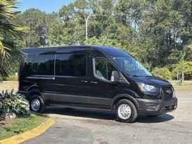 A black 2025 Ford Transit 350 van with large windows and a sleek design is parked, showcasing its modern front grille and spacious build