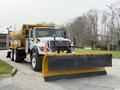 A 2016 International 7600 truck equipped with a yellow snow plow at the front and a large material hopper in the back