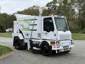 A white 2012 Global M4 street sweeper with green and yellow graphics featuring a front-mounted broom and a compact design for urban cleaning