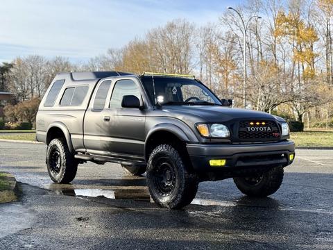 A black 2004 Toyota Tacoma with larger tires and a raised suspension features a cap on the bed and a light bar on the roof