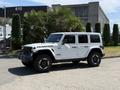 A white 2021 Jeep Wrangler Rubicon with black wheels and a distinctive exterior design is parked with a greenery backdrop