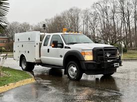 A white 2015 Ford F-450 truck with a utility body and yellow emergency lights on the roof is parked on a wet street