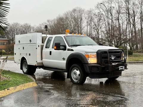 A white 2015 Ford F-450 truck with a utility body and yellow emergency lights on the roof is parked on a wet street