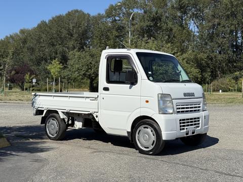 A white 2009 Suzuki Carry truck with a flatbed body facing slightly to the right parked on a gravel surface