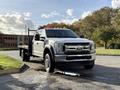 A 2019 Ford F-550 pickup truck with a flatbed and silver exterior is parked with its lights on