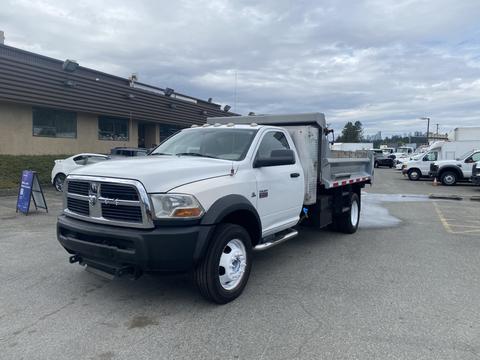 A 2011 Dodge Ram 5500 truck in white with a flatbed and chrome accents parked in a lot with a visible water spray on the ground
