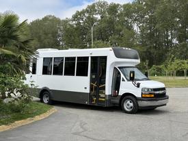 A white 2020 Chevrolet Express shuttle bus with large windows and yellow interior seating is parked with its door open