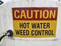 A sign on a trailer reads CAUTION HOT WATER WEED CONTROL with a connector for a hose attached to the side