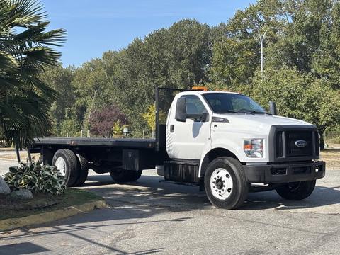 A white 2018 Ford F-750 flatbed truck with a black grille and orange light bar on the roof parked with a flatbed at the rear